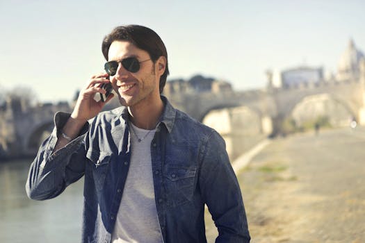 一个戴着太阳镜的时尚男子，在阳光明媚的日子里，正微笑着在河边用智能手机聊天