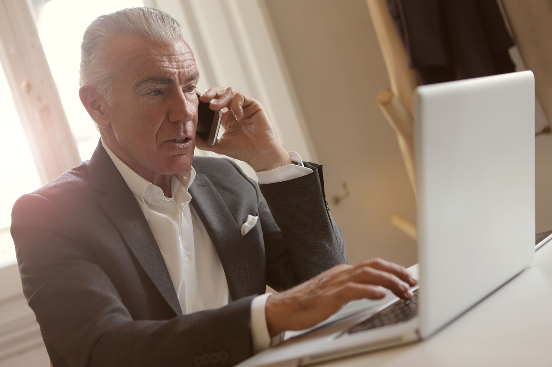 Senior businessman multitasking with a phone call while working on a laptop in a modern office setting.
