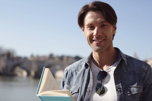 Man In Blue Denim Jacket Smiling While Holding A Book