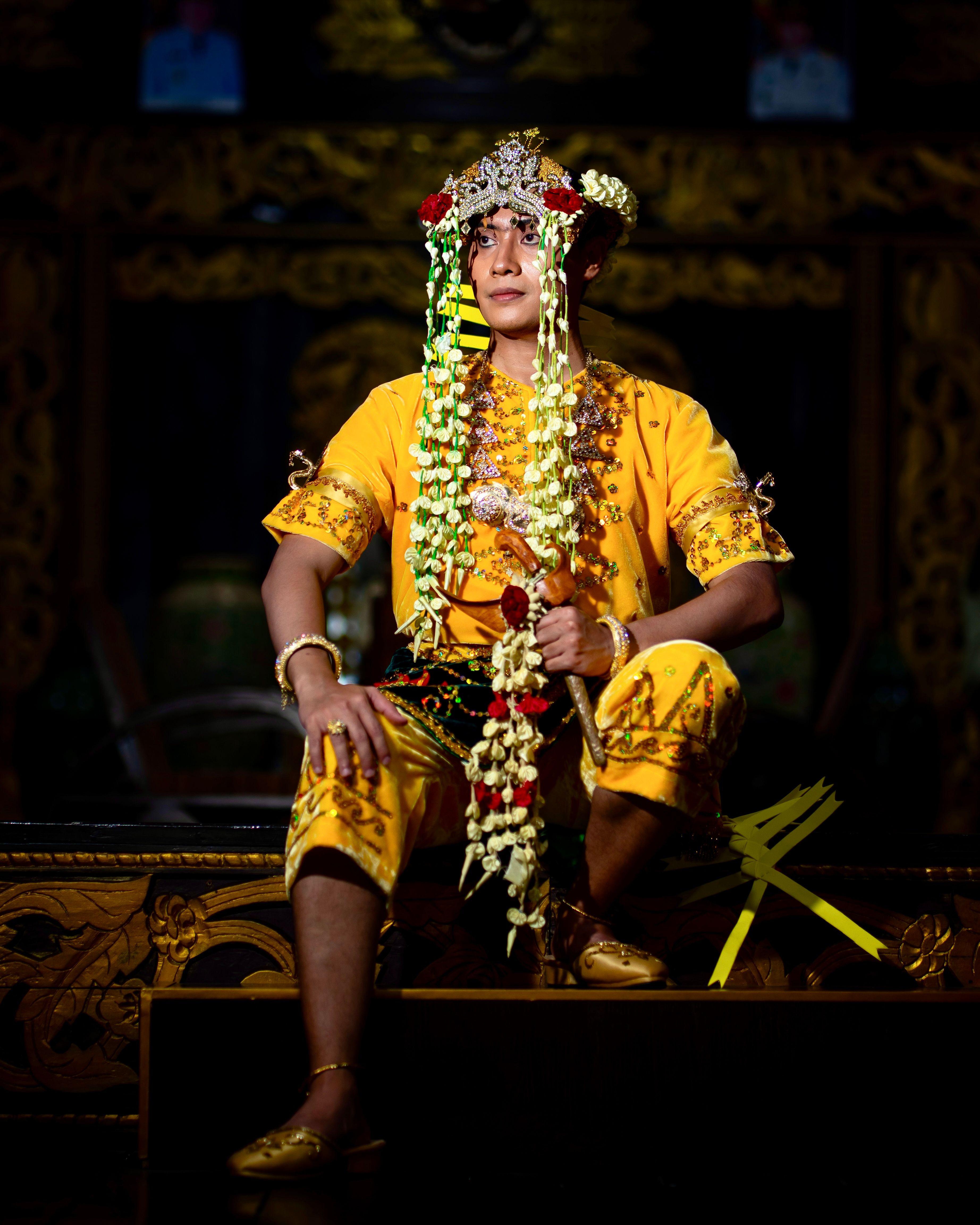 A person wearing traditional Balinese attire and headpiece sitting on a stage in dramatic lighting.