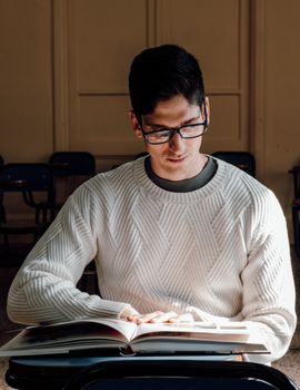 Male student in white sweater reads a book in a Buenos Aires university classroom, focused on learning.
