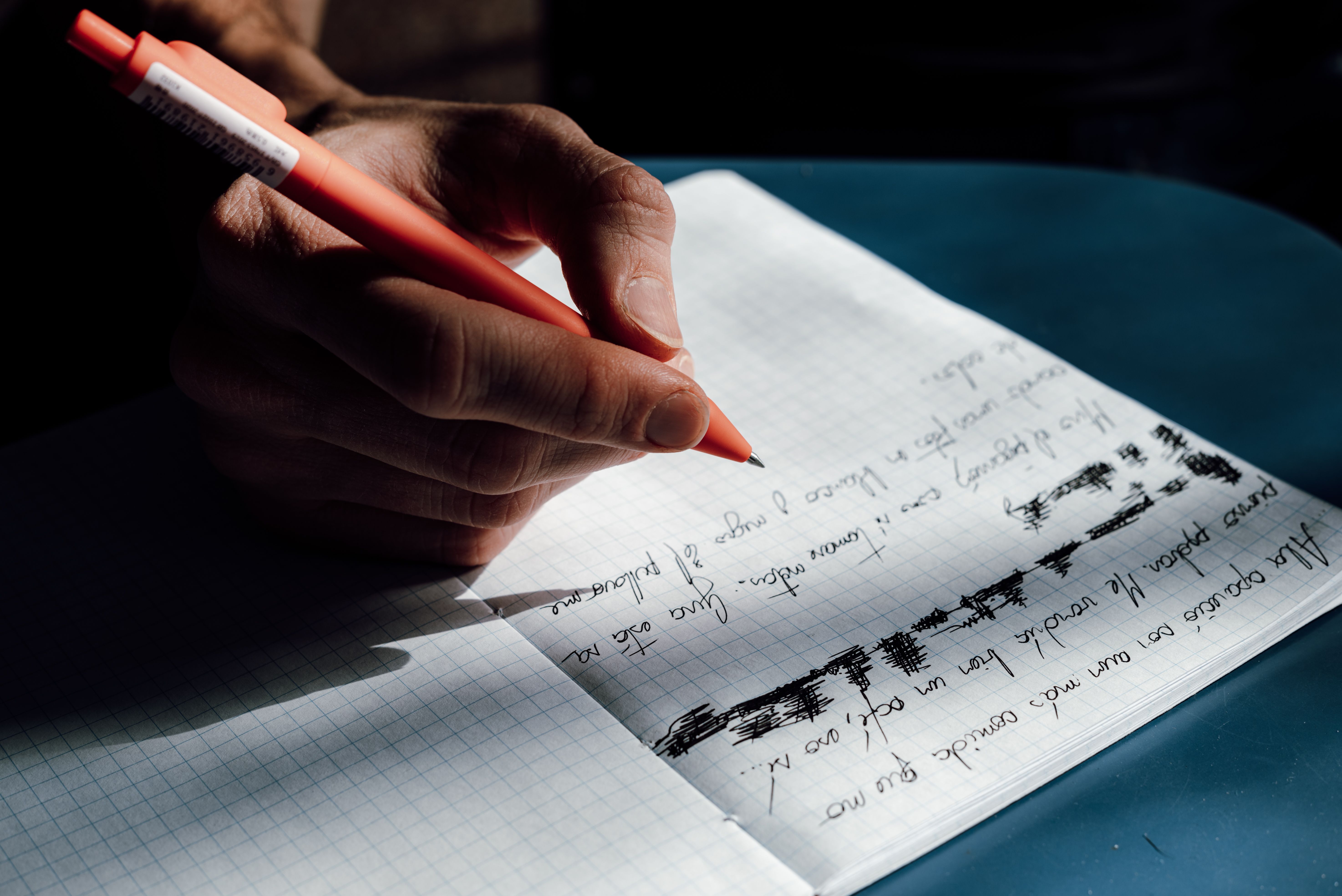 A hand writing with a red pen on a grid notebook, capturing thoughtful detail.
