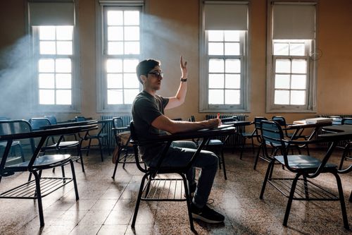 Free A student raises hand in a university classroom, Buenos Aires, featuring warm light and empty desks. Stock Photo