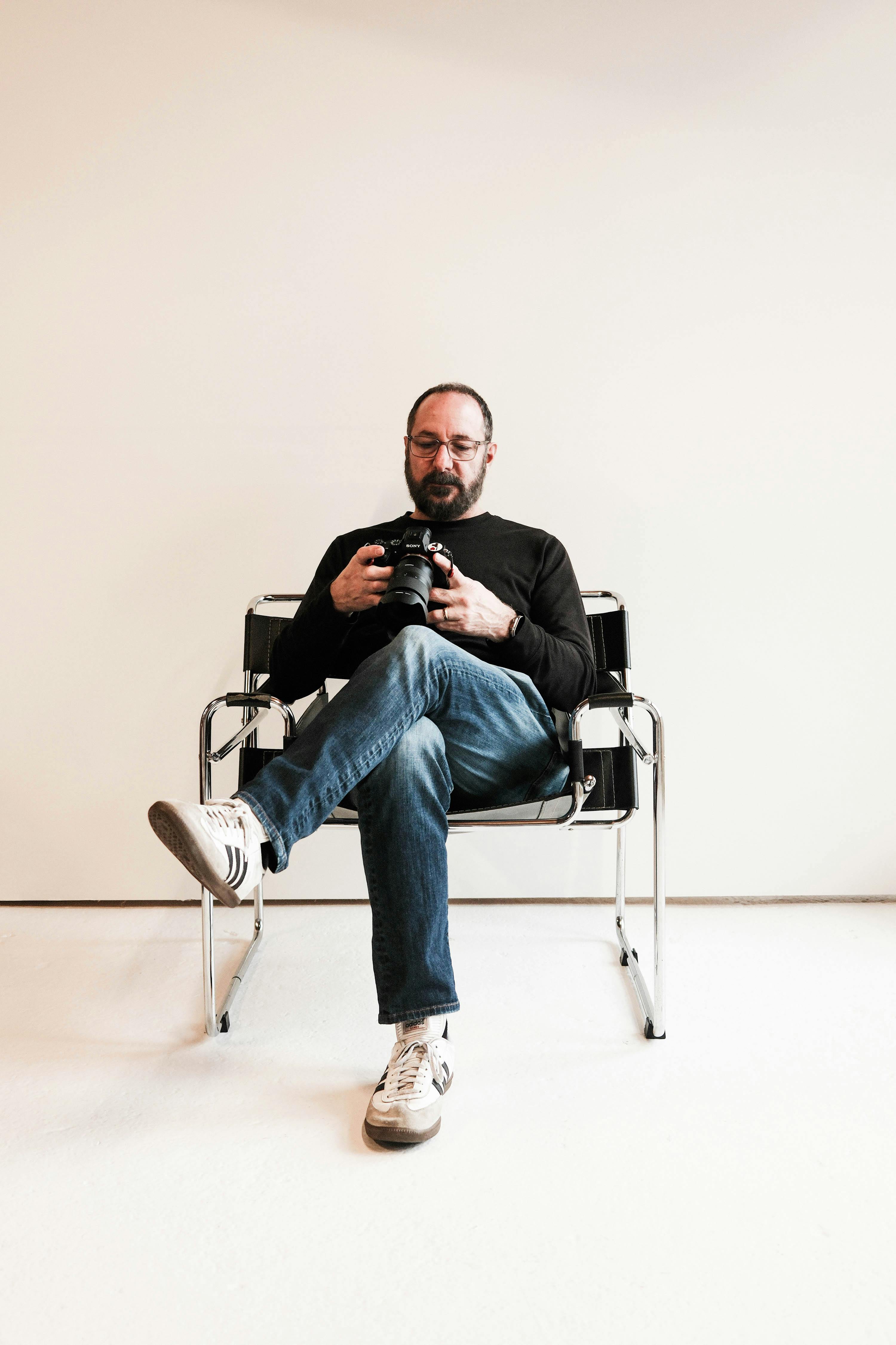 A man with a camera sits casually in a modern chair against a neutral backdrop.