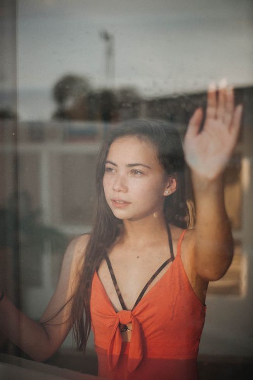 Free A young woman in an orange top gazes thoughtfully out a window, hand raised against the glass. Stock Photo