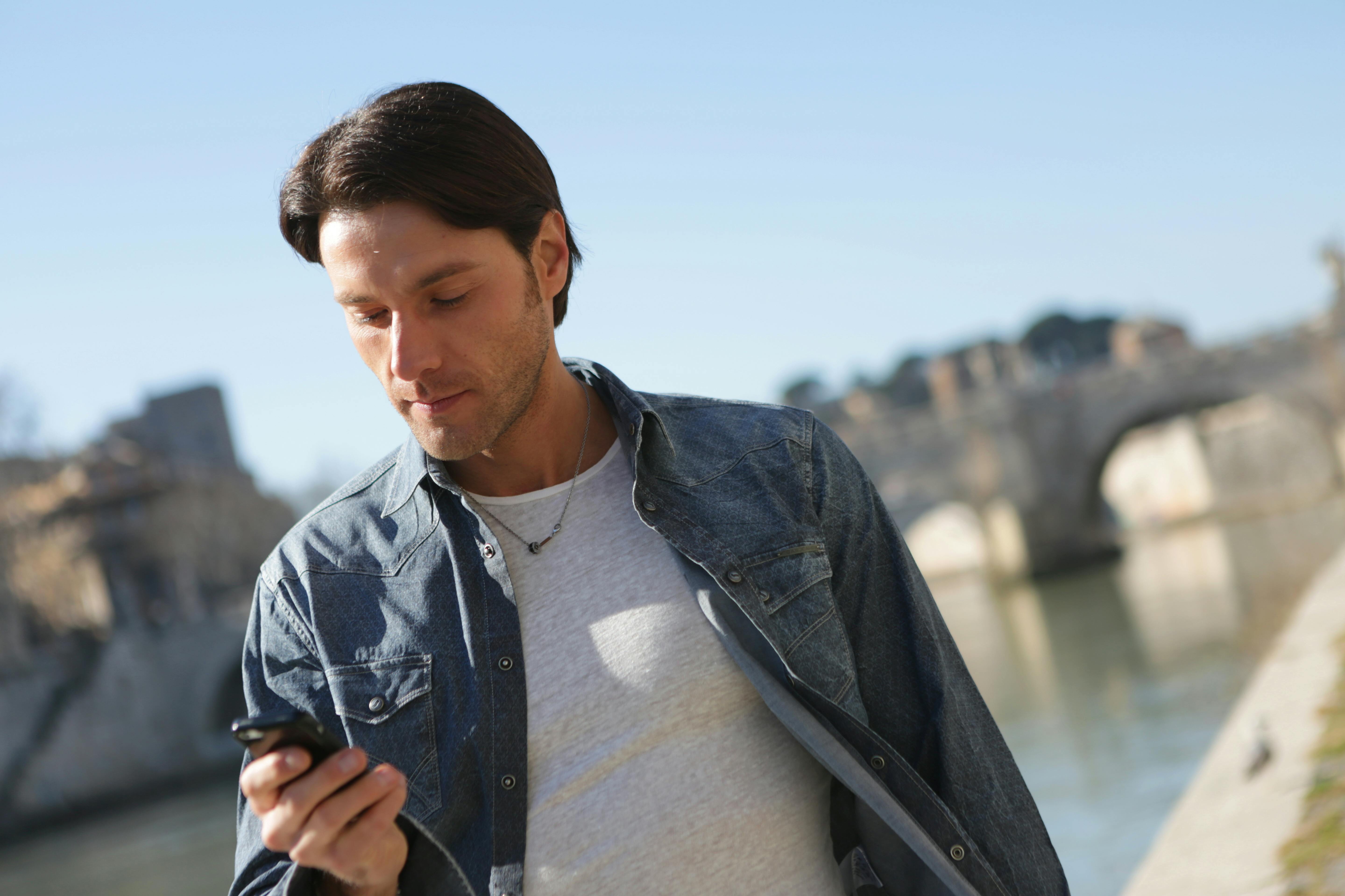 一名穿着牛仔夹克的时尚男子在跨河的桥边查看手机