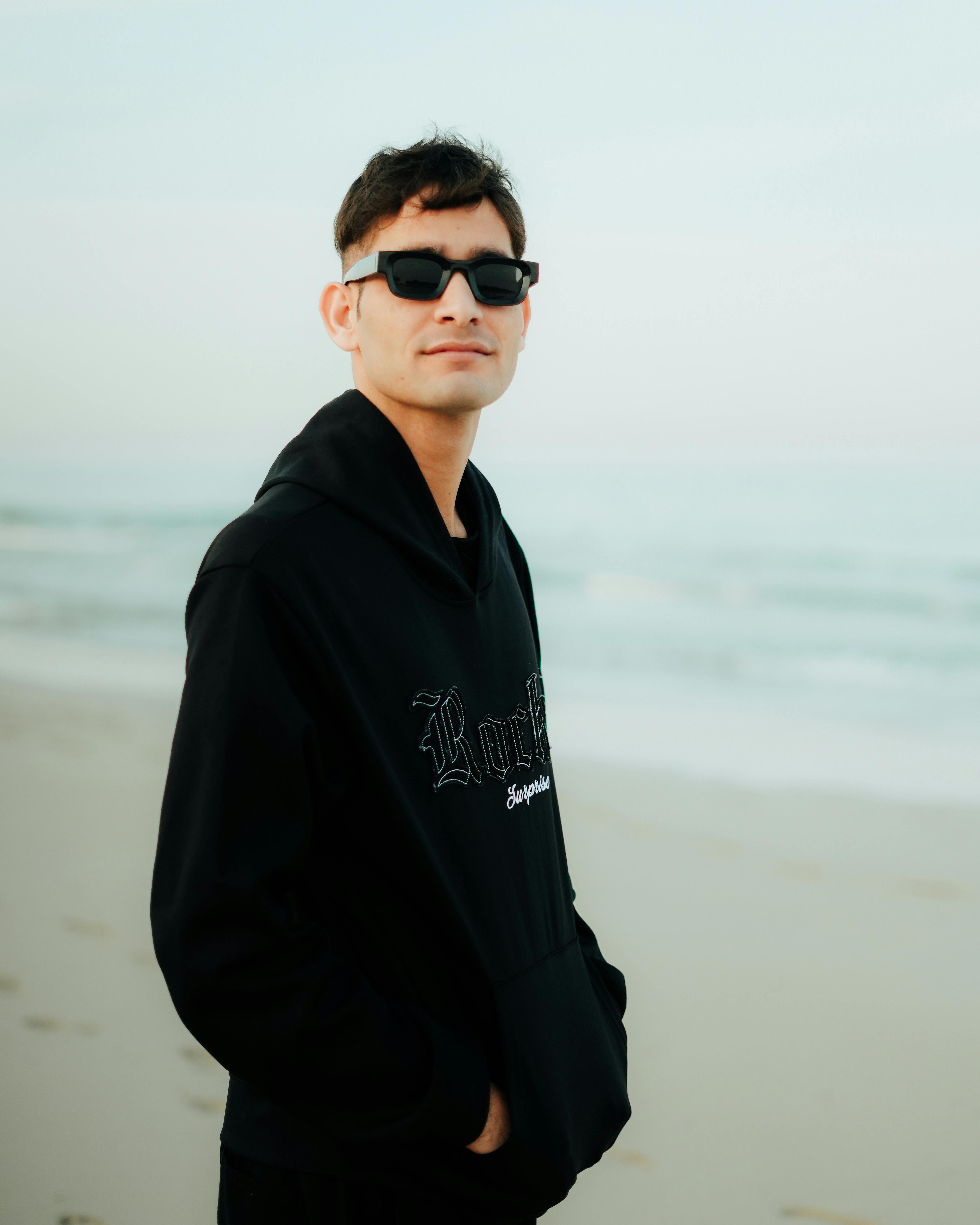 Young man in black hoodie and sunglasses enjoying a stroll on a Saudi Arabian beach.