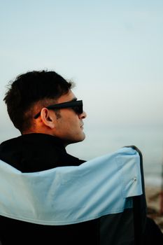 A man wearing sunglasses sitting relaxed on a chair by the sea in Saudi Arabia during the day.