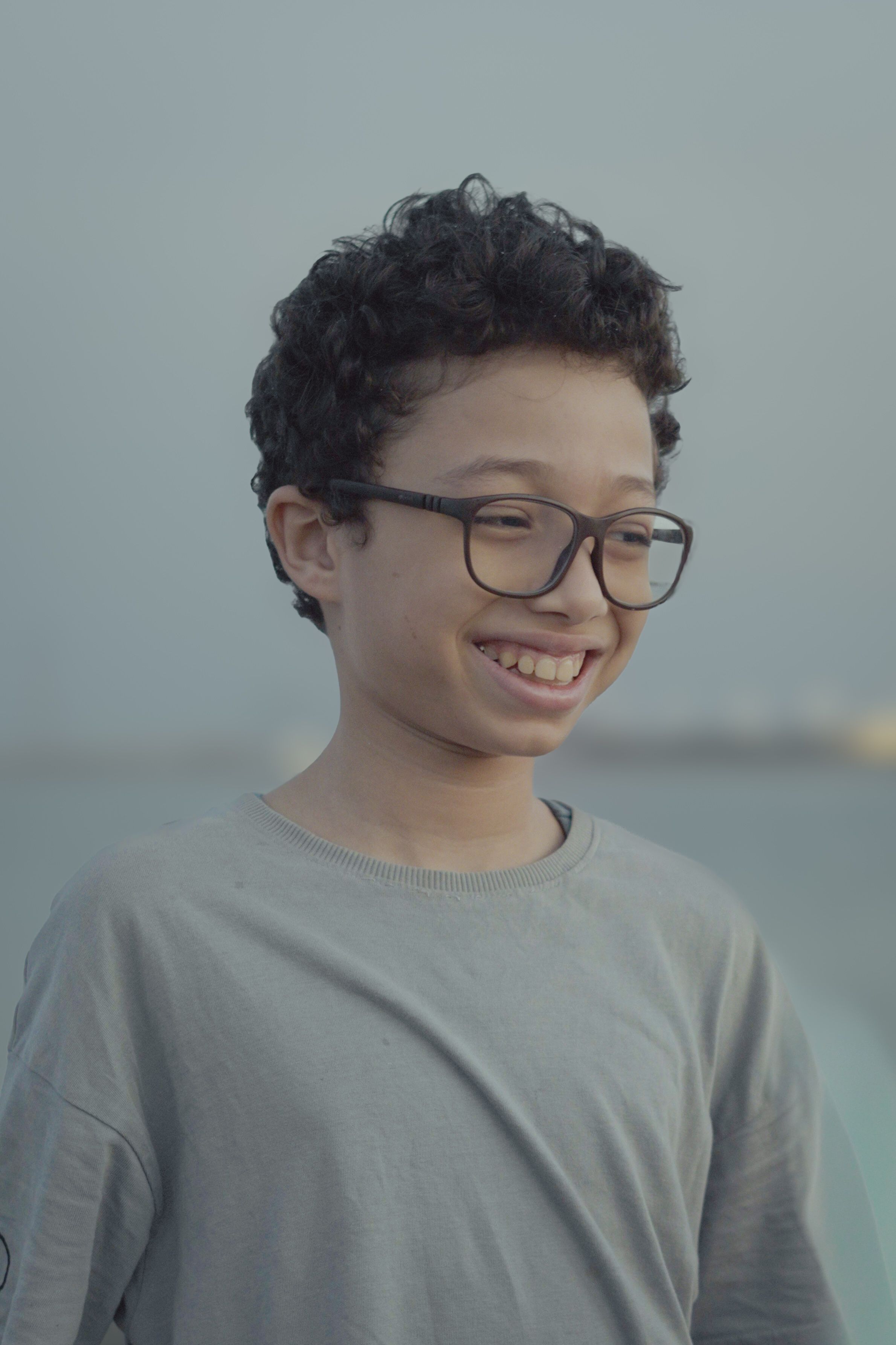 A young boy smiling while standing by the waterfront in Saudi Arabia with a serene background.