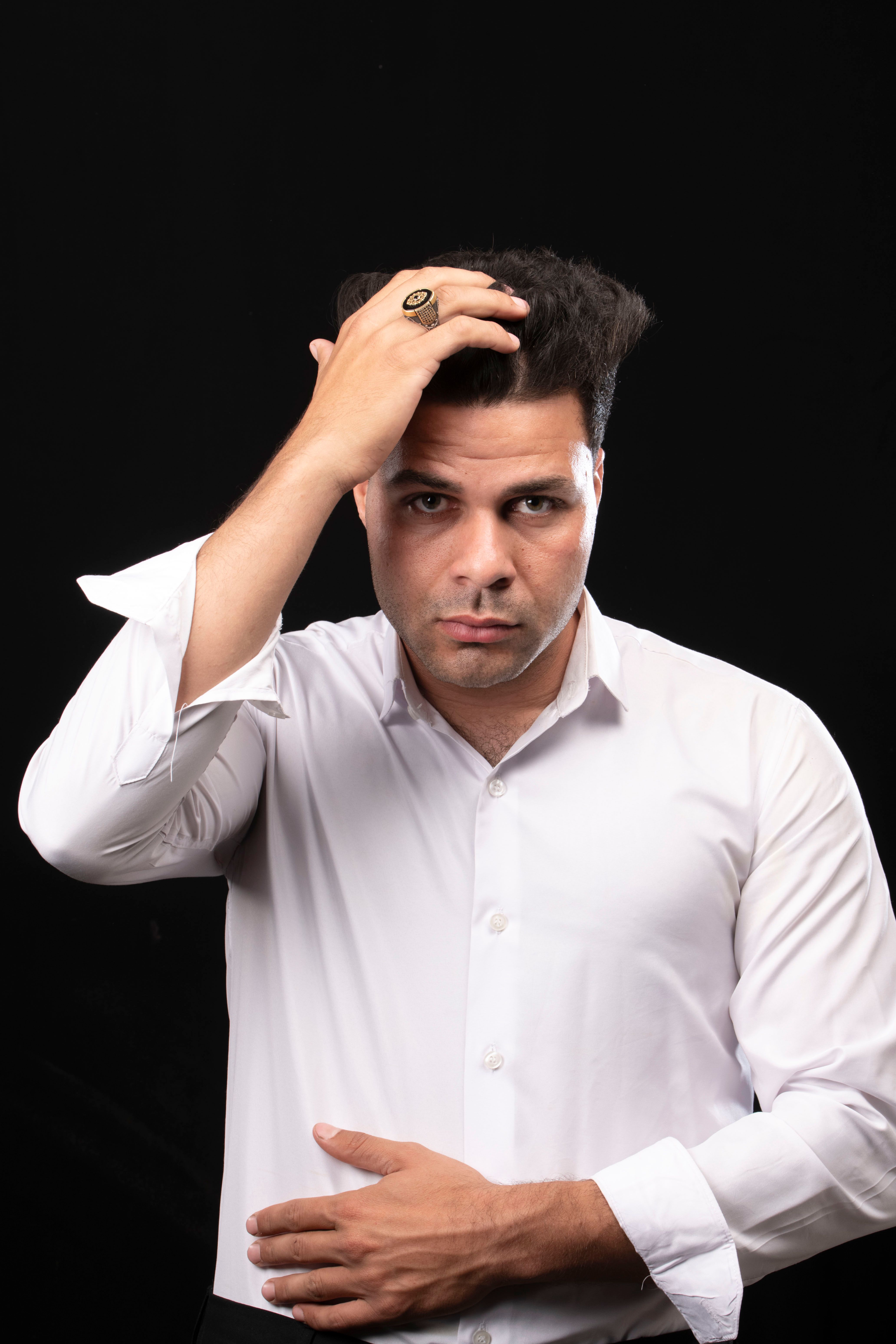Studio portrait of a man in a white shirt adjusting his hair against a black background.