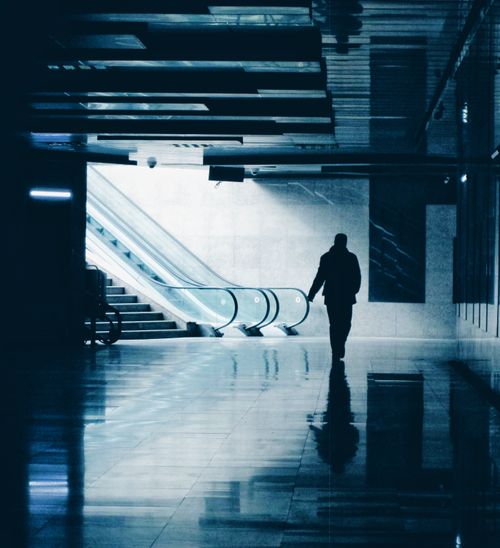 Free Silhouette of a solitary figure walking through a modern indoor setting with an escalator, casting reflections on a glossy floor. Stock Photo