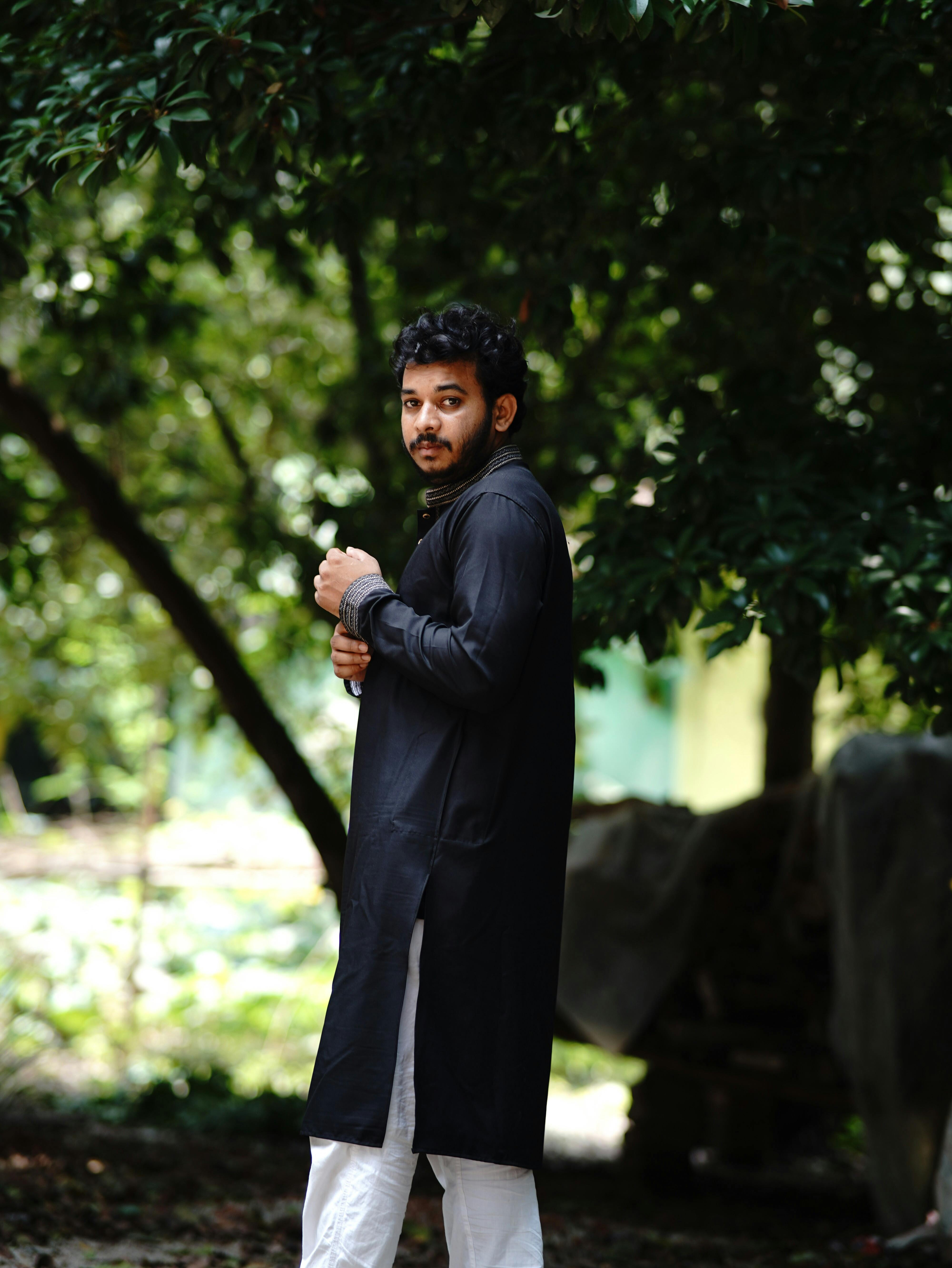A young man stands outdoors in traditional attire, surrounded by lush greenery.