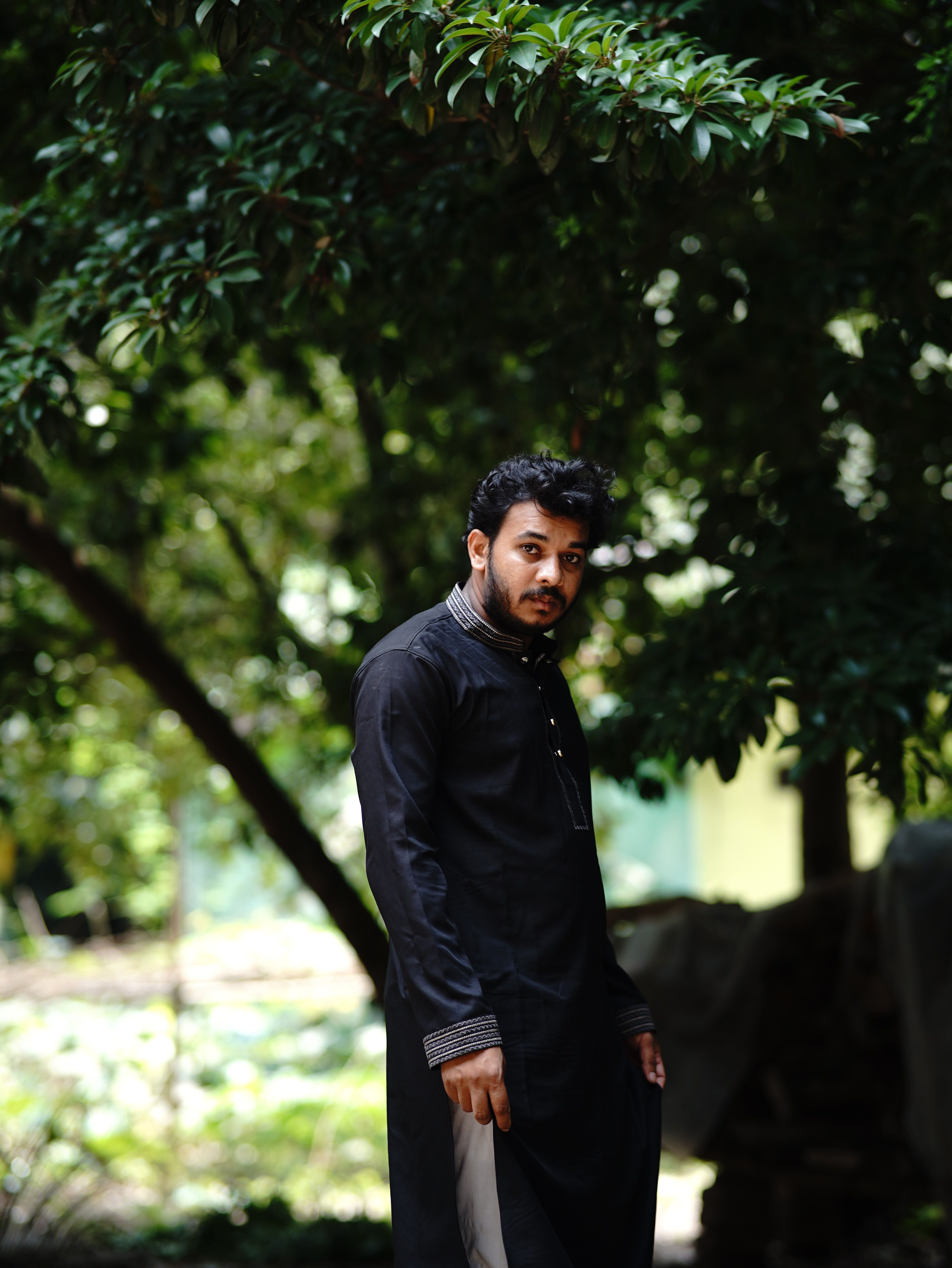 A thoughtful young man standing outdoors in Dhaka, Bangladesh under lush green trees.