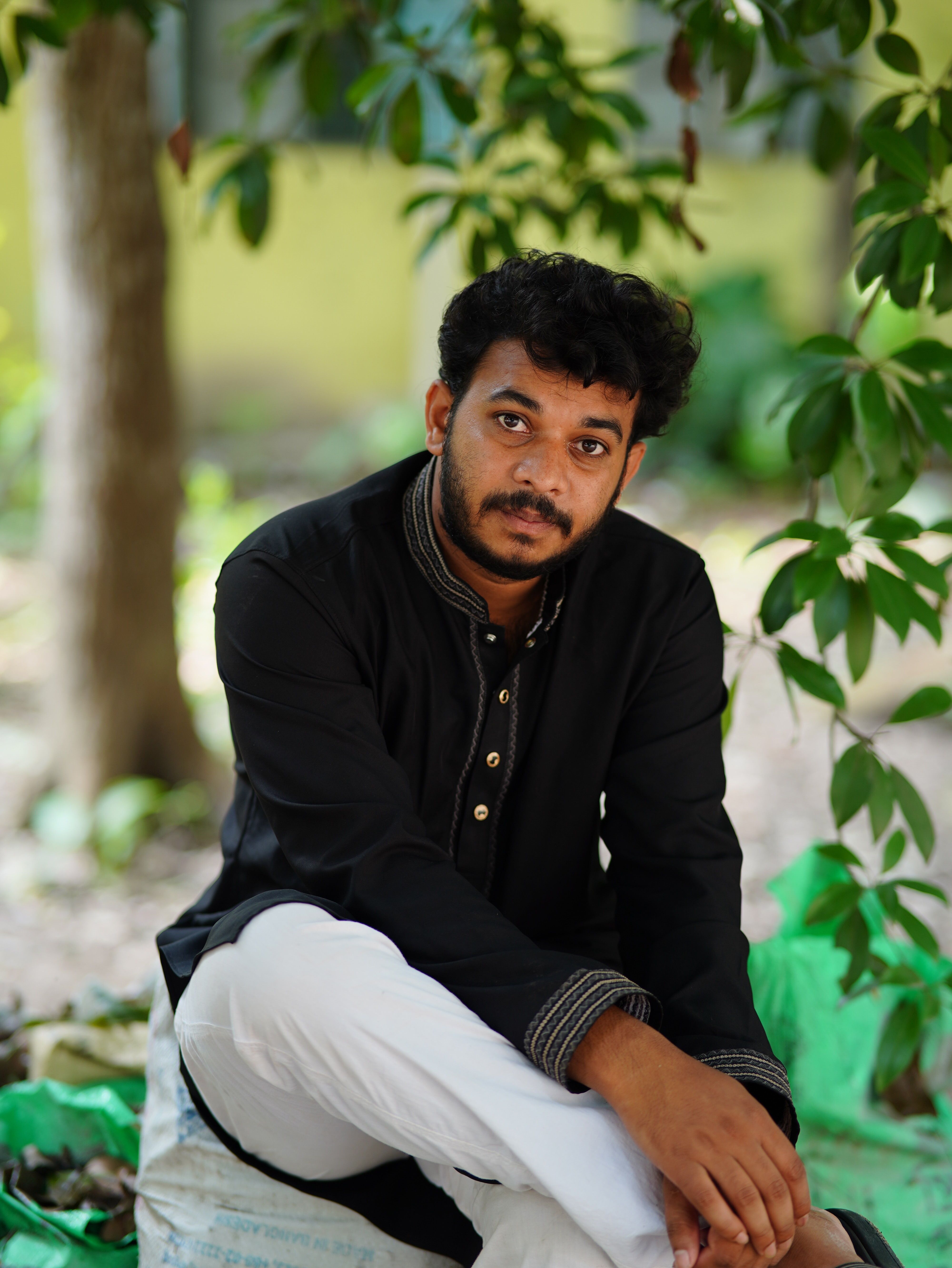 Portrait of a young man sitting outdoors in traditional attire, surrounded by greenery.