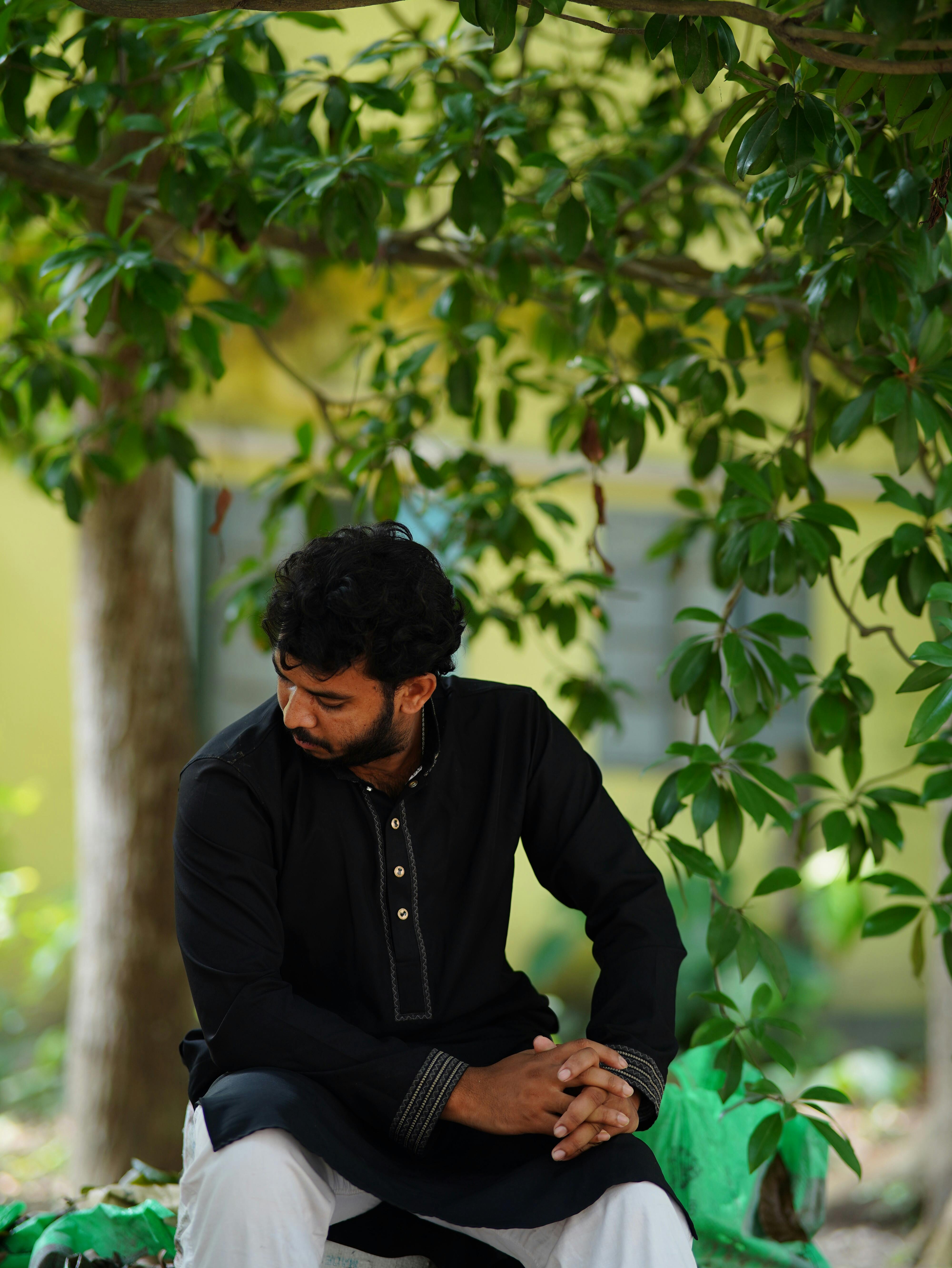 South Asian man in traditional black attire sitting under lush foliage, outdoors in Bangladesh.