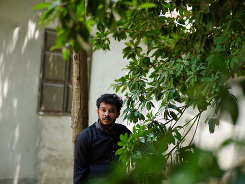 Free A man in traditional attire stands outdoors beside lush green foliage. Stock Photo
