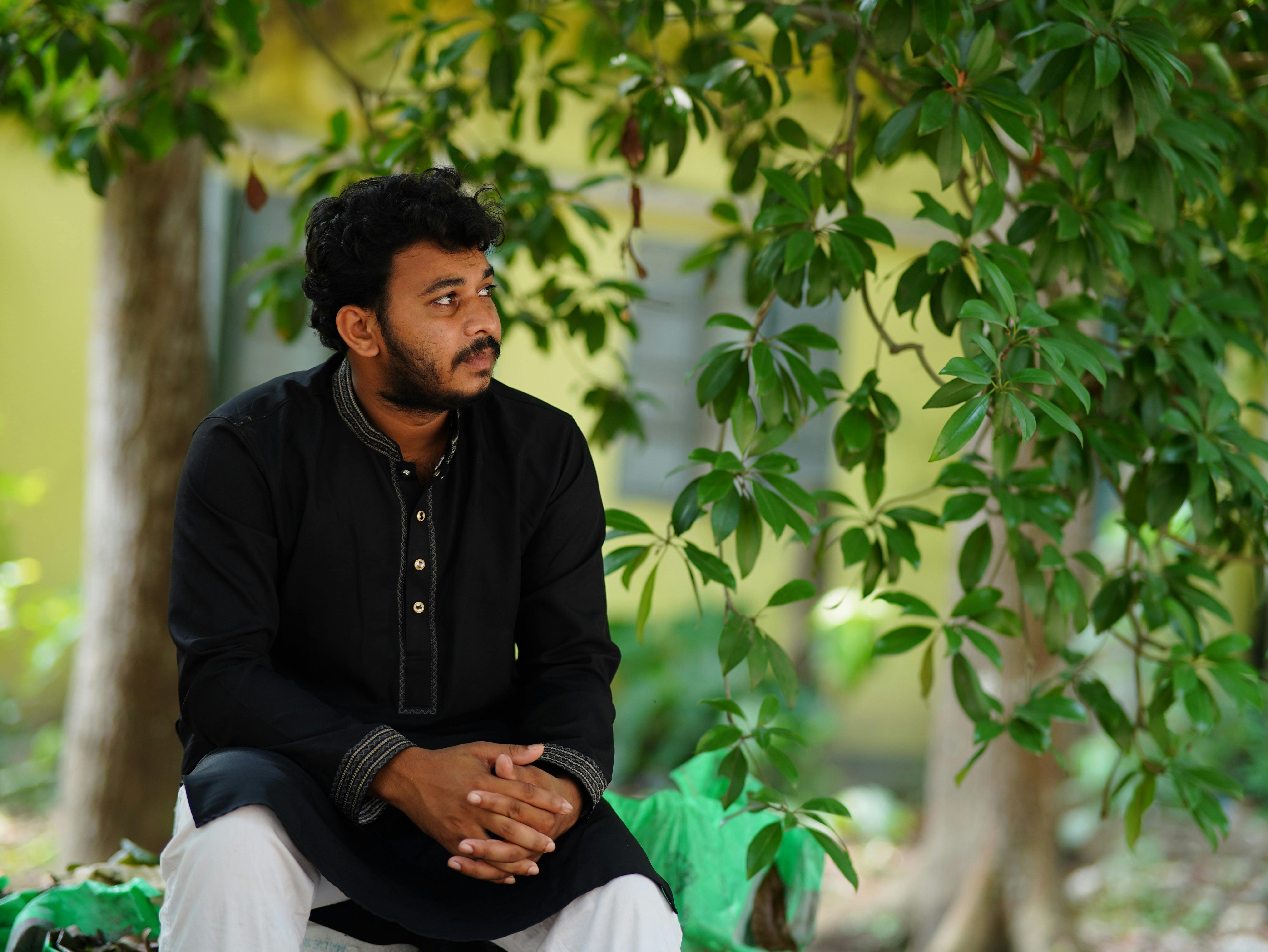 A man in traditional attire sitting outdoors surrounded by greenery, capturing a moment of peaceful reflection.