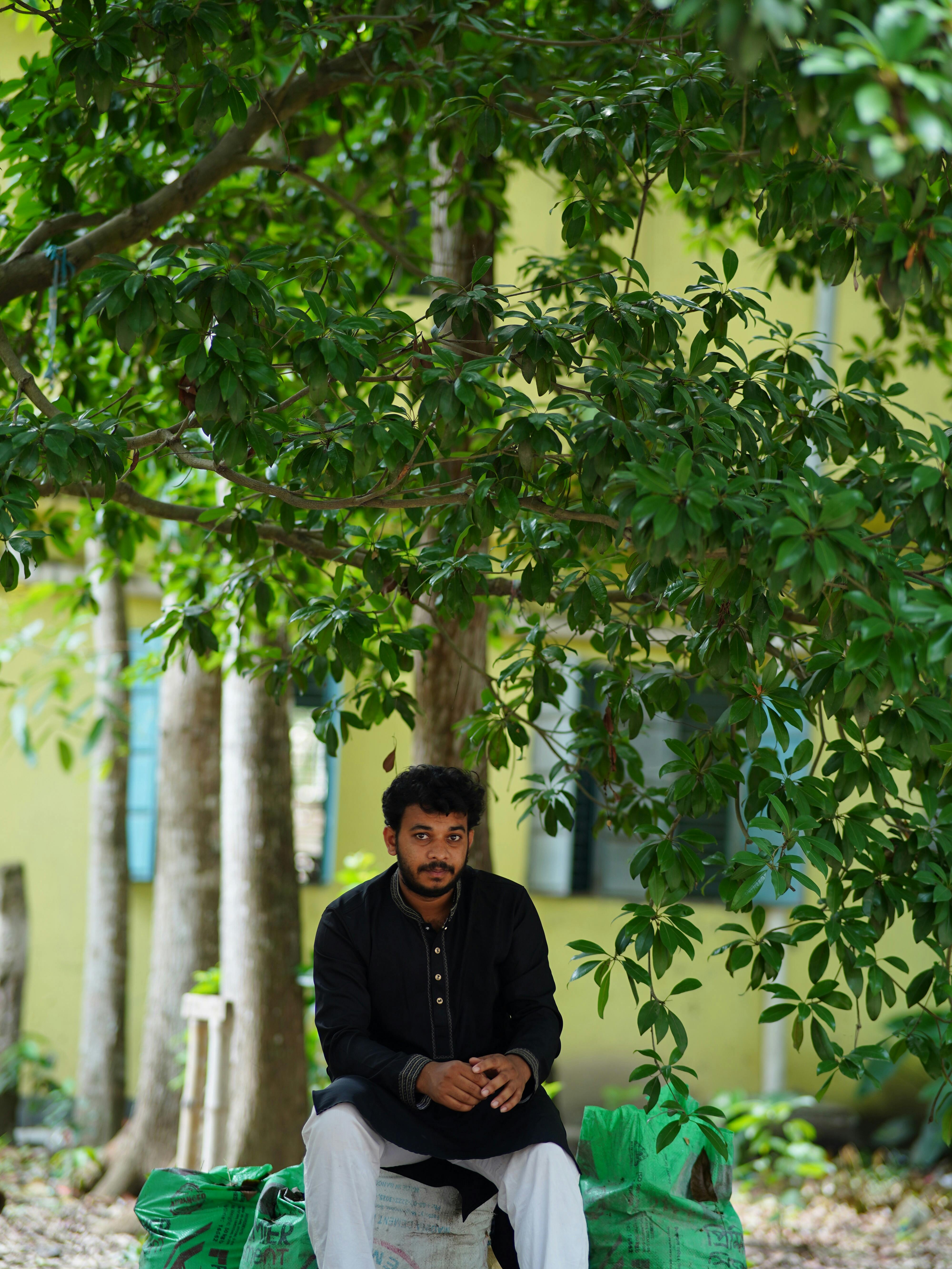 A man in traditional attire sits peacefully under lush trees in Dhaka, Bangladesh.