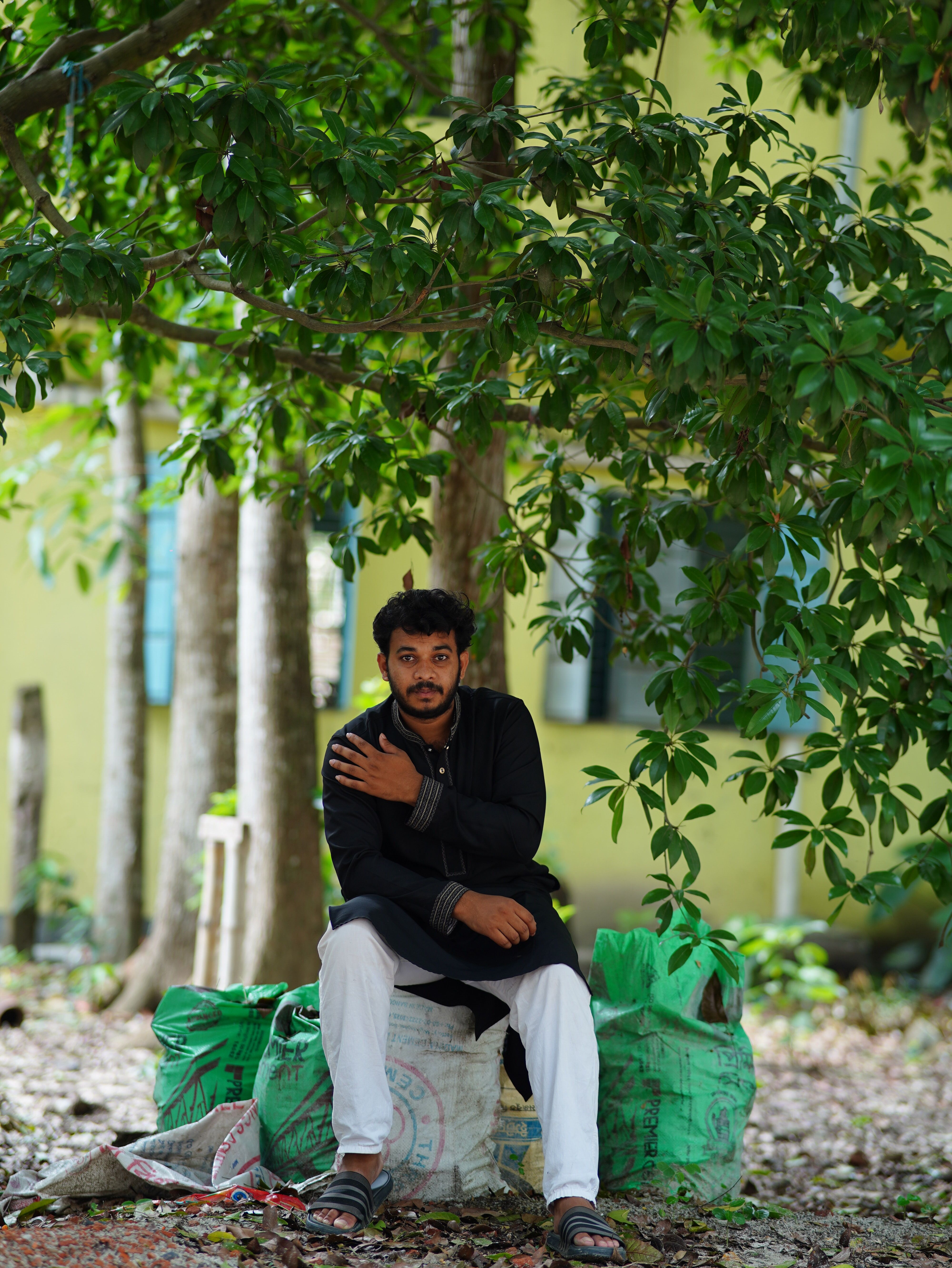 Young man sitting outdoors under a tree in Dhaka, Bangladesh, surrounded by nature.