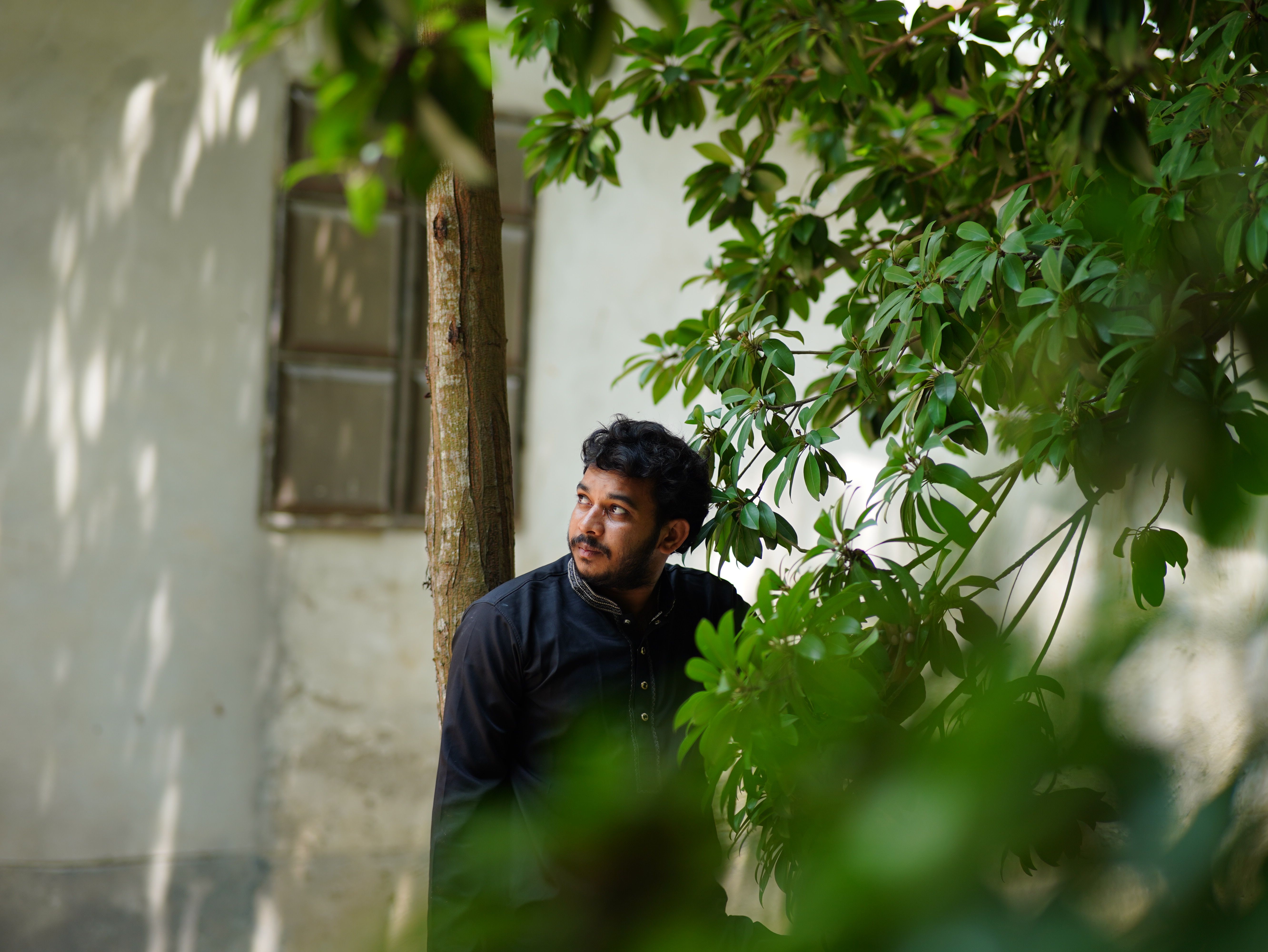 A young man in traditional attire stands outdoors surrounded by greenery.