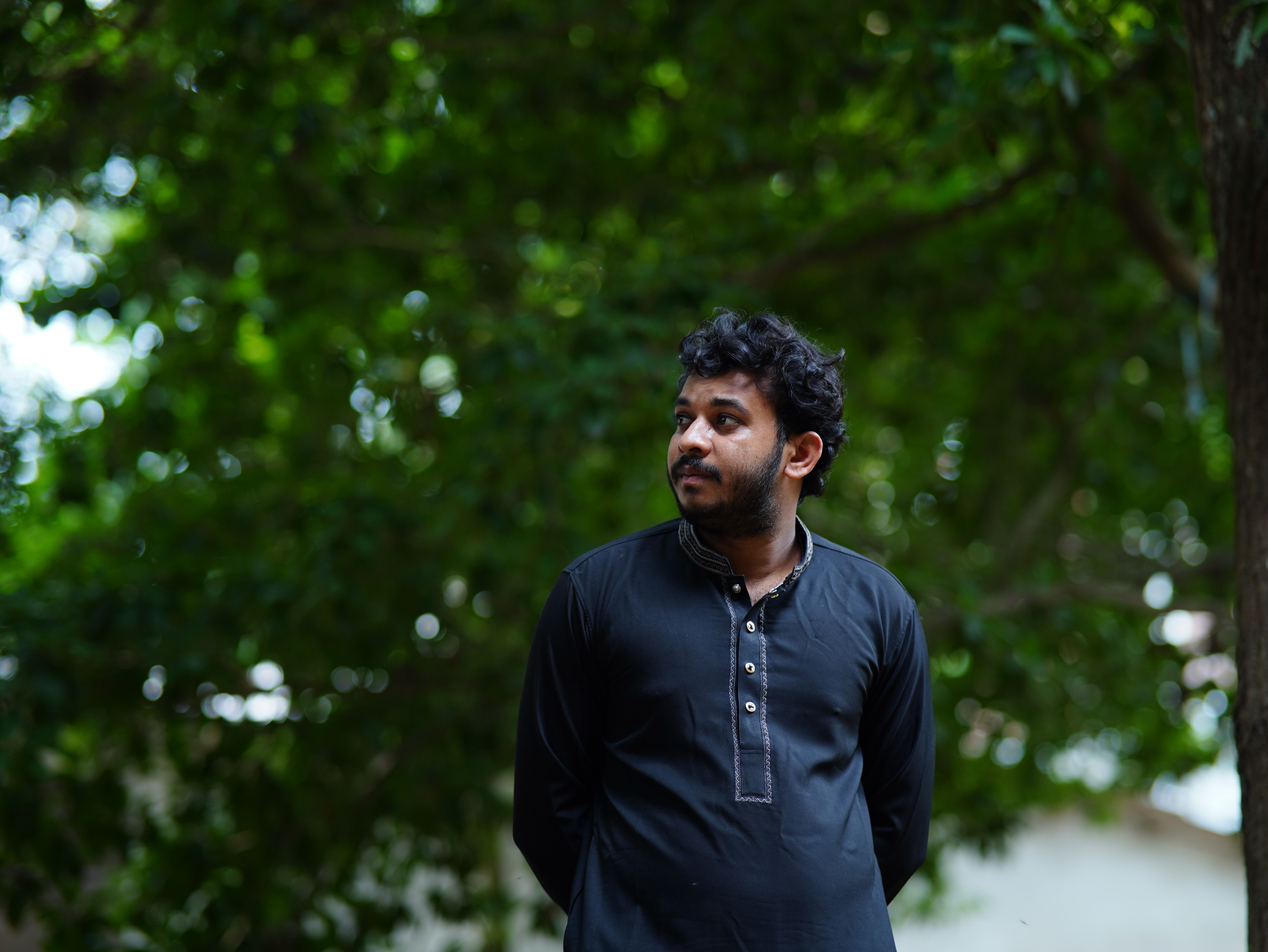 A man in traditional black attire looks thoughtful outdoors in a leafy setting.