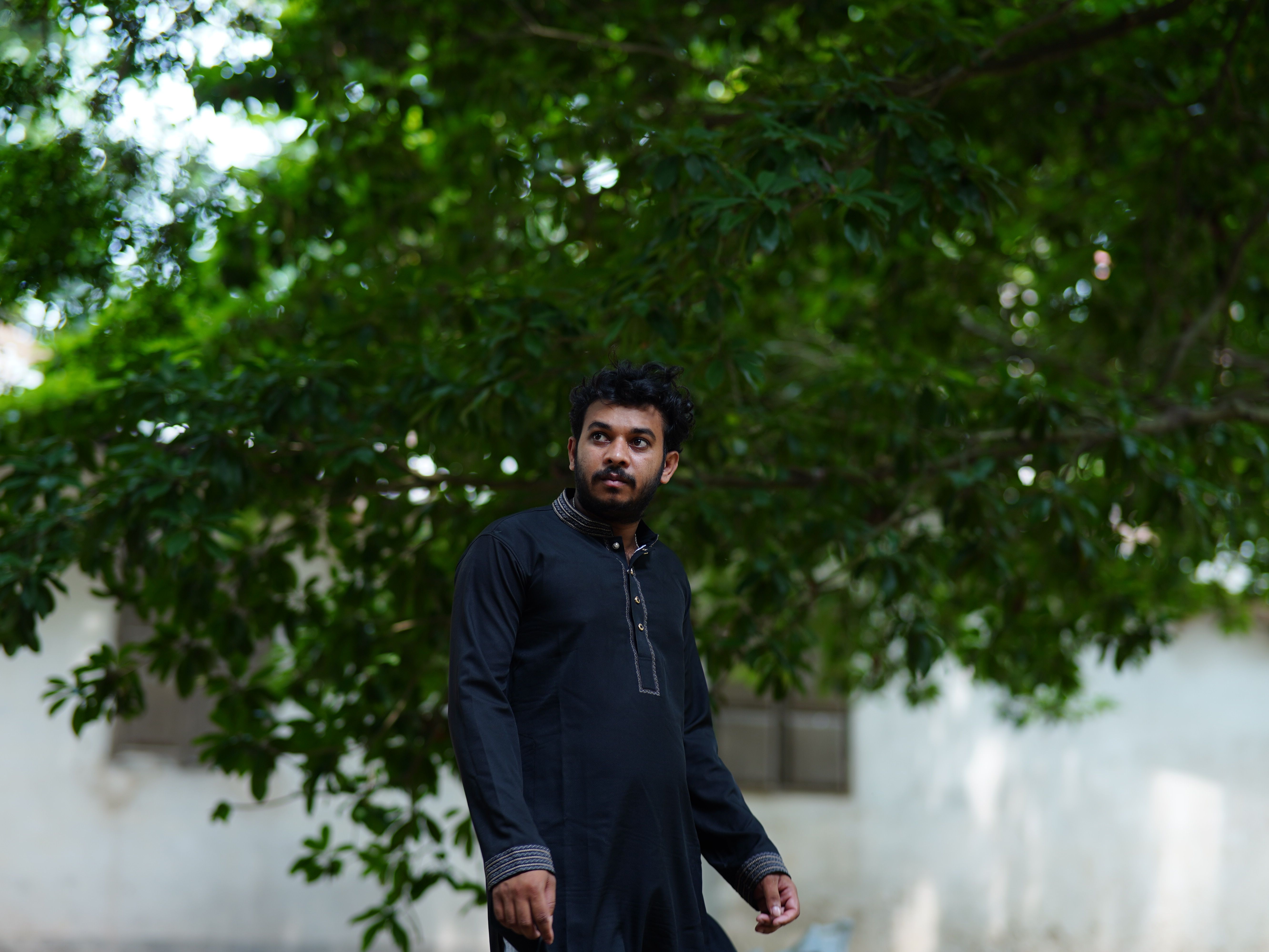A man wearing traditional black attire stands outdoors under a lush green tree.
