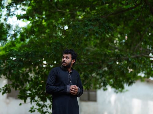 Free South Asian man standing outdoors in traditional black outfit surrounded by green trees. Stock Photo