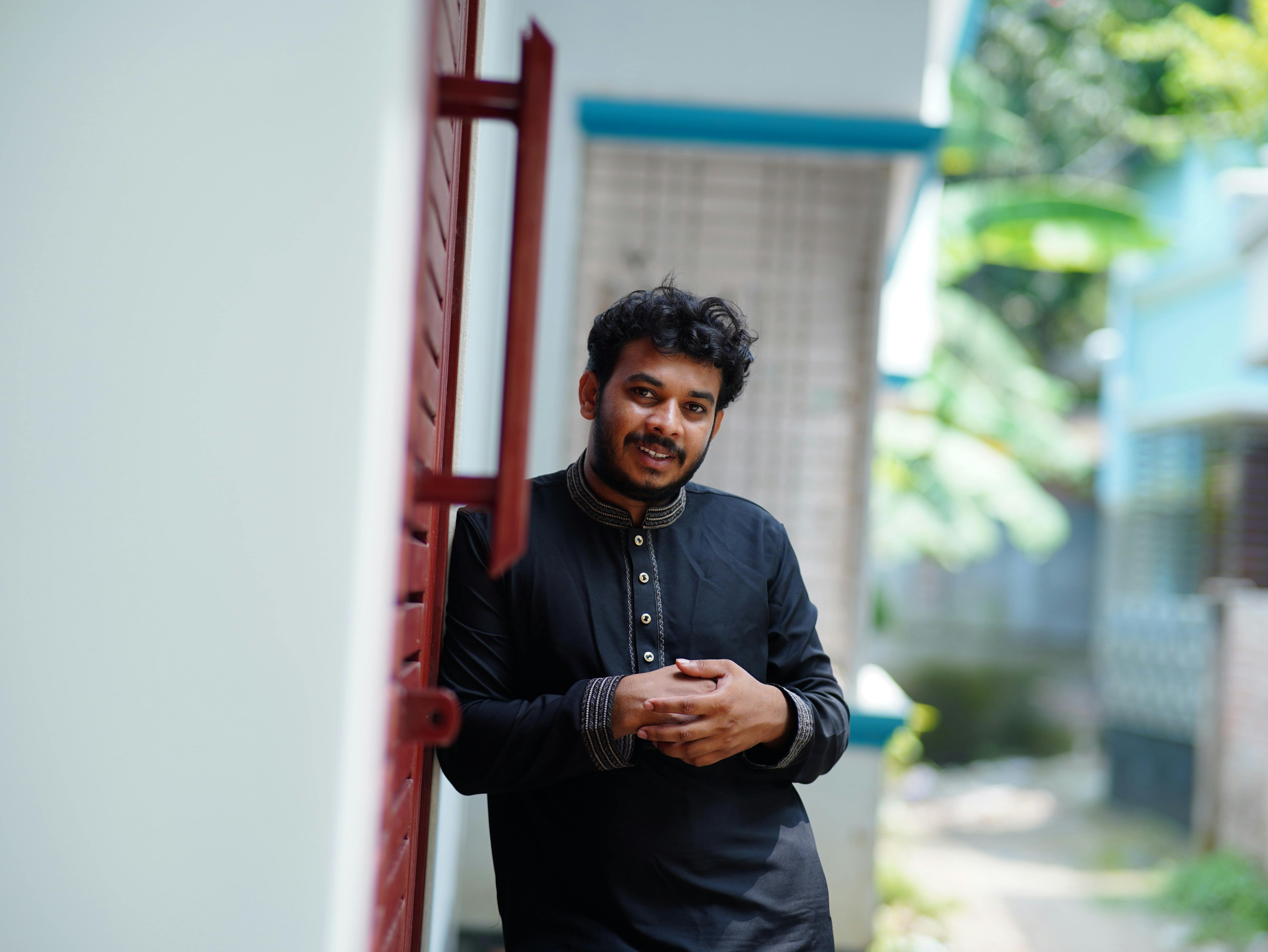 Smiling man in traditional outfit leaning against a wall outdoors in Dhaka, Bangladesh.