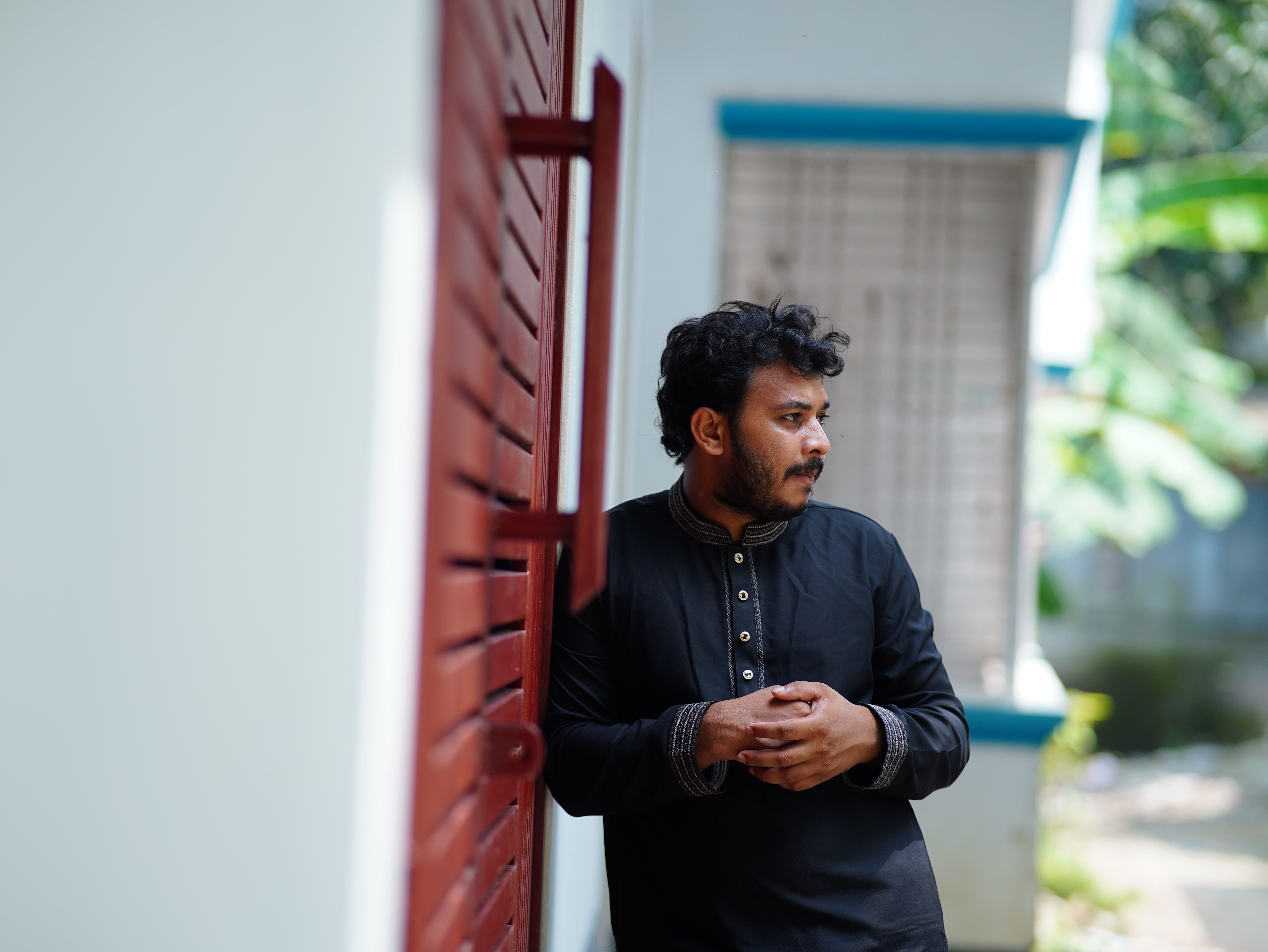 A South Asian man in traditional attire leans against a red shutter, captured in a candid pose outdoors.