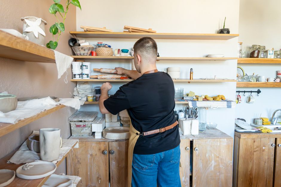 A ceramic artist in an apron organizing shelves in a cozy pottery workshop.