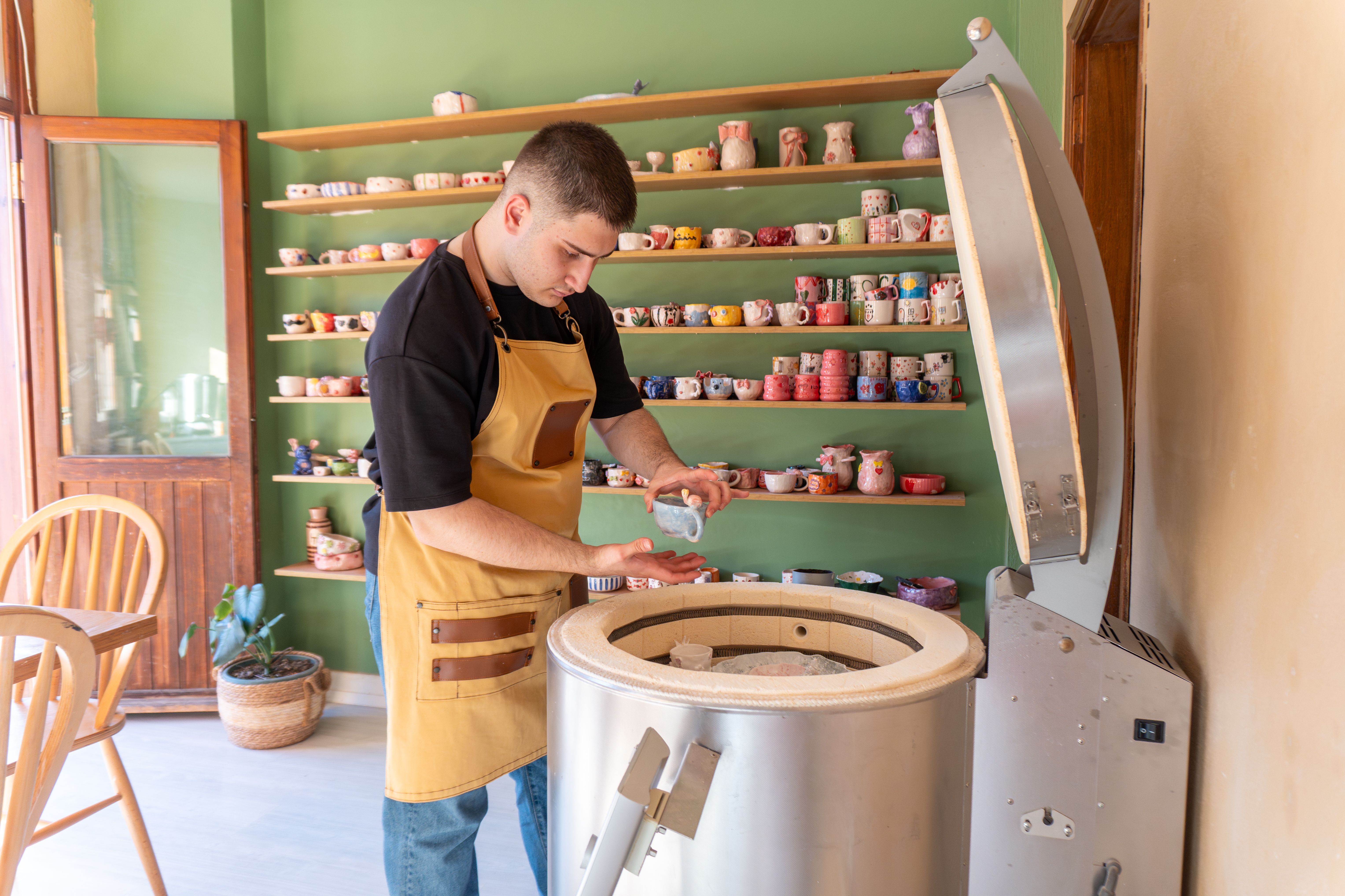 A potter prepares clay for firing in a kiln, surrounded by colorful ceramics in a well-lit studio.