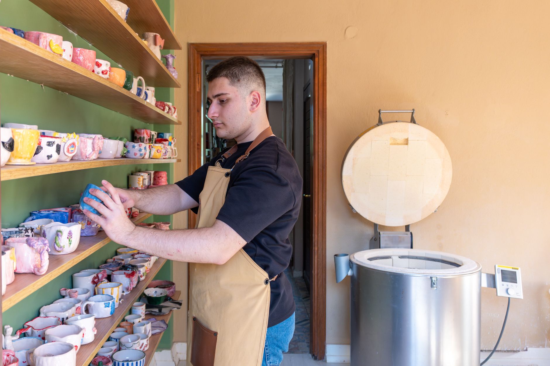 Young male potter organizing colorful handmade ceramic mugs in a pottery studio with a kiln.