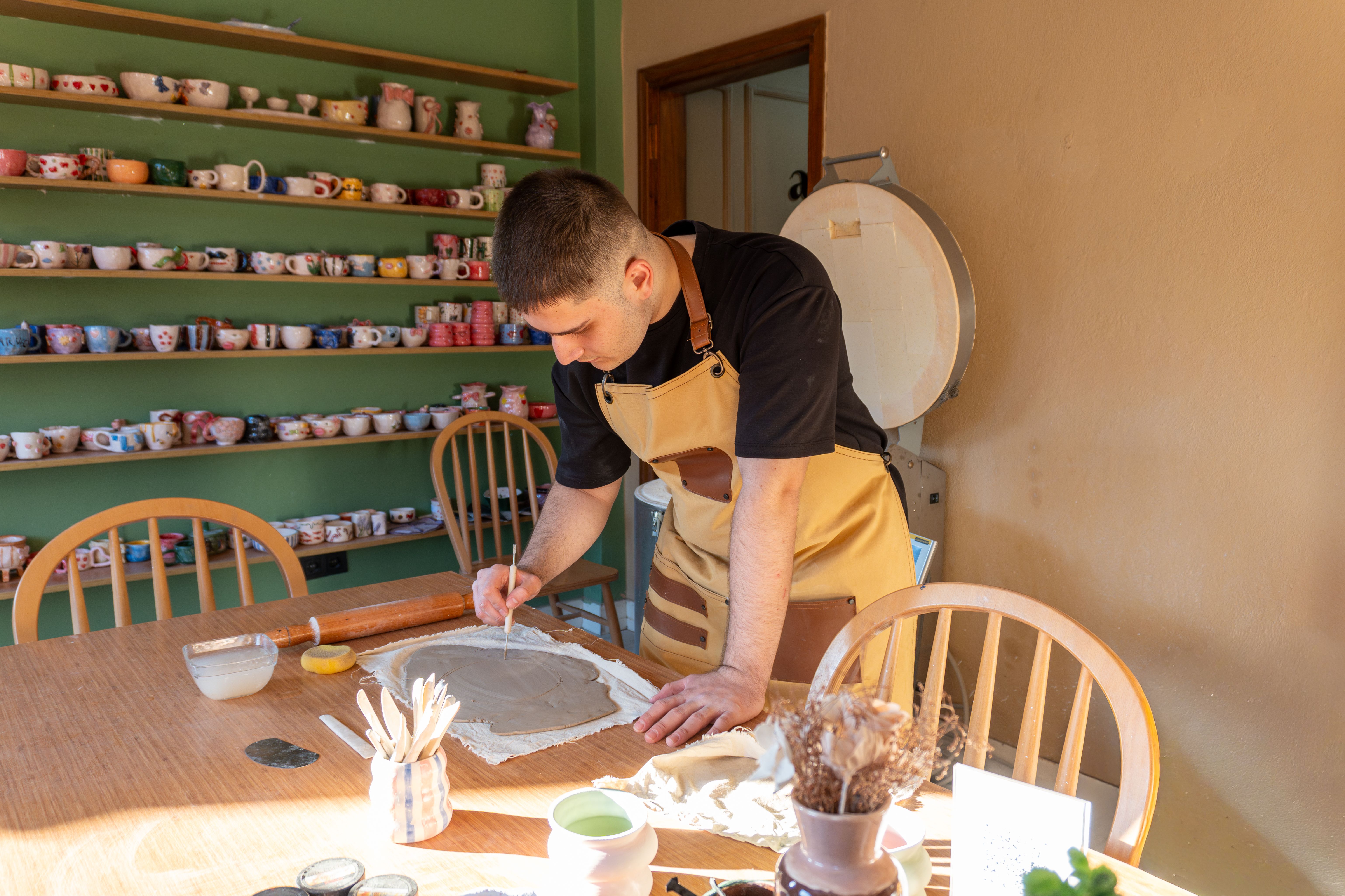 A potter working at a sunlit bench, shelves of unfired pots behind him