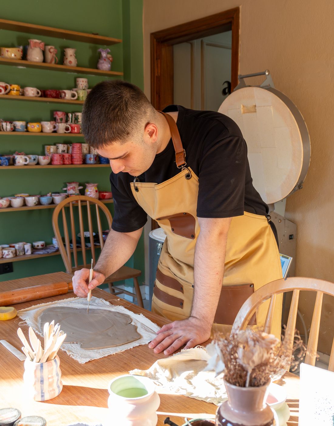 A potter working at a sunlit bench, shelves of unfired pots behind him