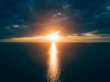 A breathtaking aerial view of the ocean at sunset with dramatic clouds and a golden horizon.