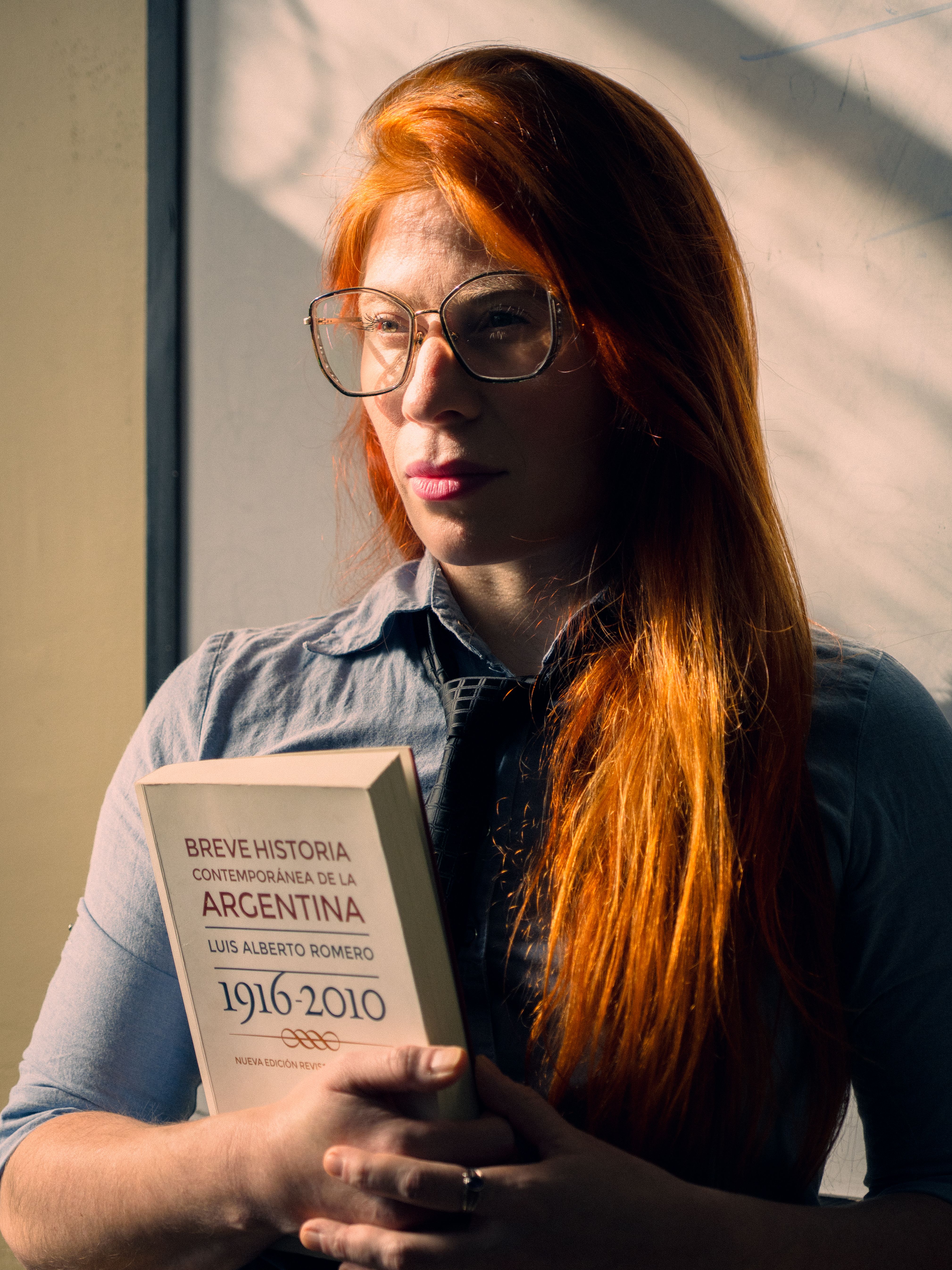 A red-haired woman holding a history book in sunlight indoors, Buenos Aires.