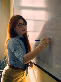 Focused teacher at a whiteboard writing lesson notes in a sunlit classroom setting.