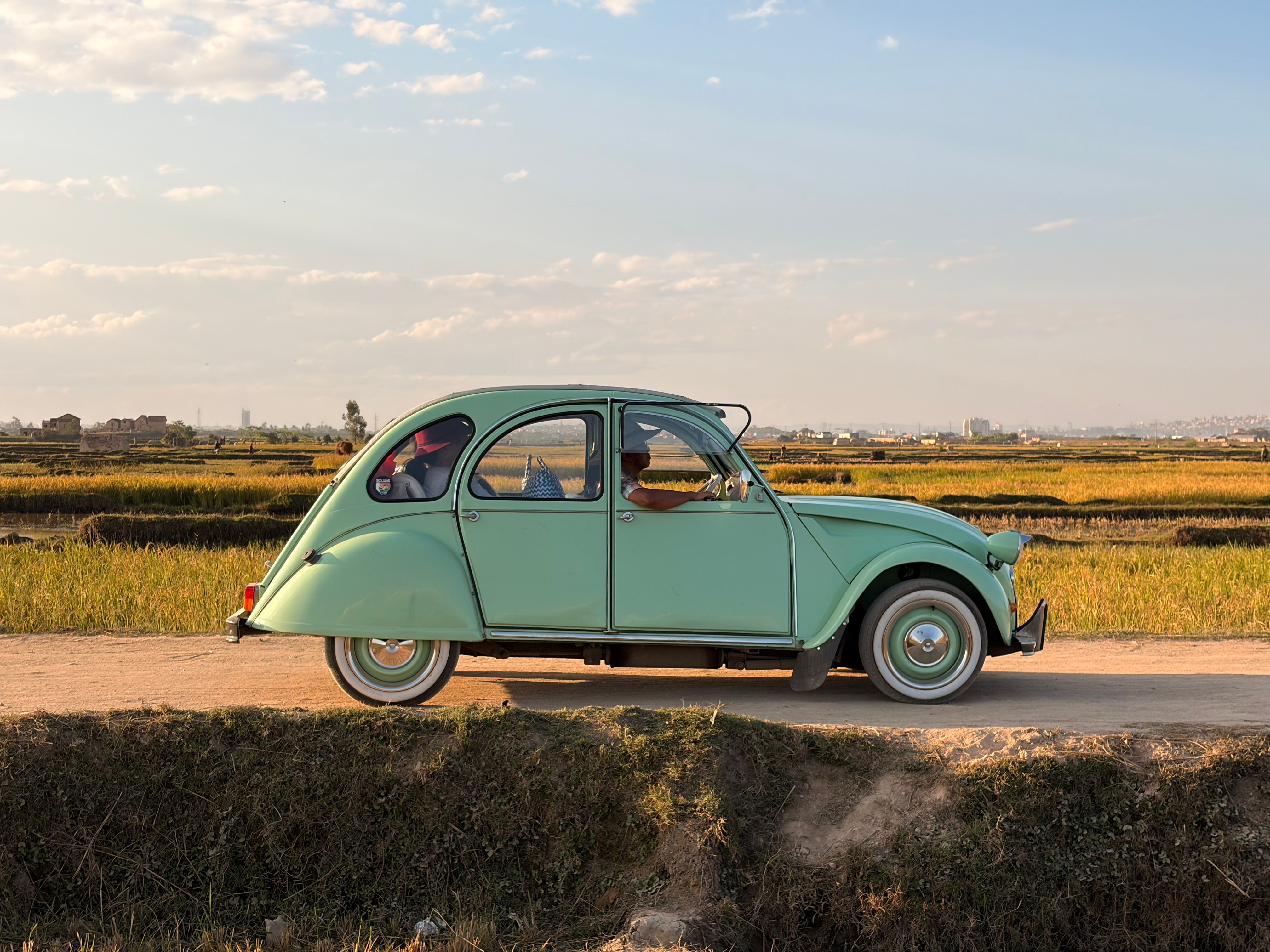 Classic mint-green car driving down a rural road in Madagascar under a sunny sky with scenic fields.