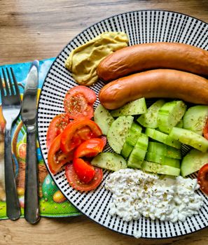 A vibrant breakfast plate featuring sausages, cucumbers, tomatoes, mustard, and cottage cheese.