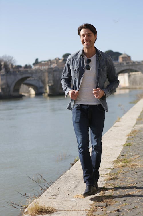 Man in Blue Shirt and Blue Denim Jeans Standing on Gray Concrete Pavement Near Body