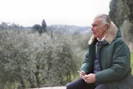 一位穿着绿色毛皮衬里夹克的老人坐在户外，手里拿着手机