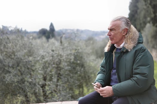 一位穿着绿色毛皮衬里夹克的老人坐在户外，手里拿着手机