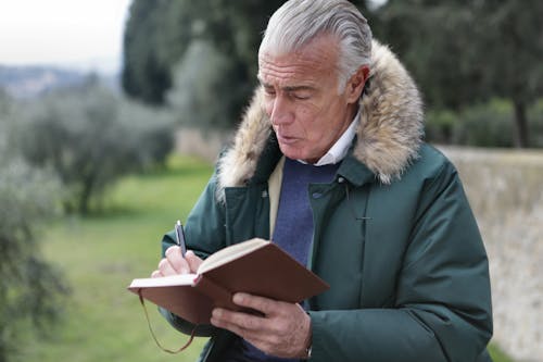 Man in Green Jacket Holding Brown Book and a Pen