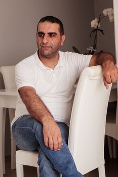 A man is casually seated indoors on a white chair, wearing a white shirt and jeans. - Fotografer Sidoarjo