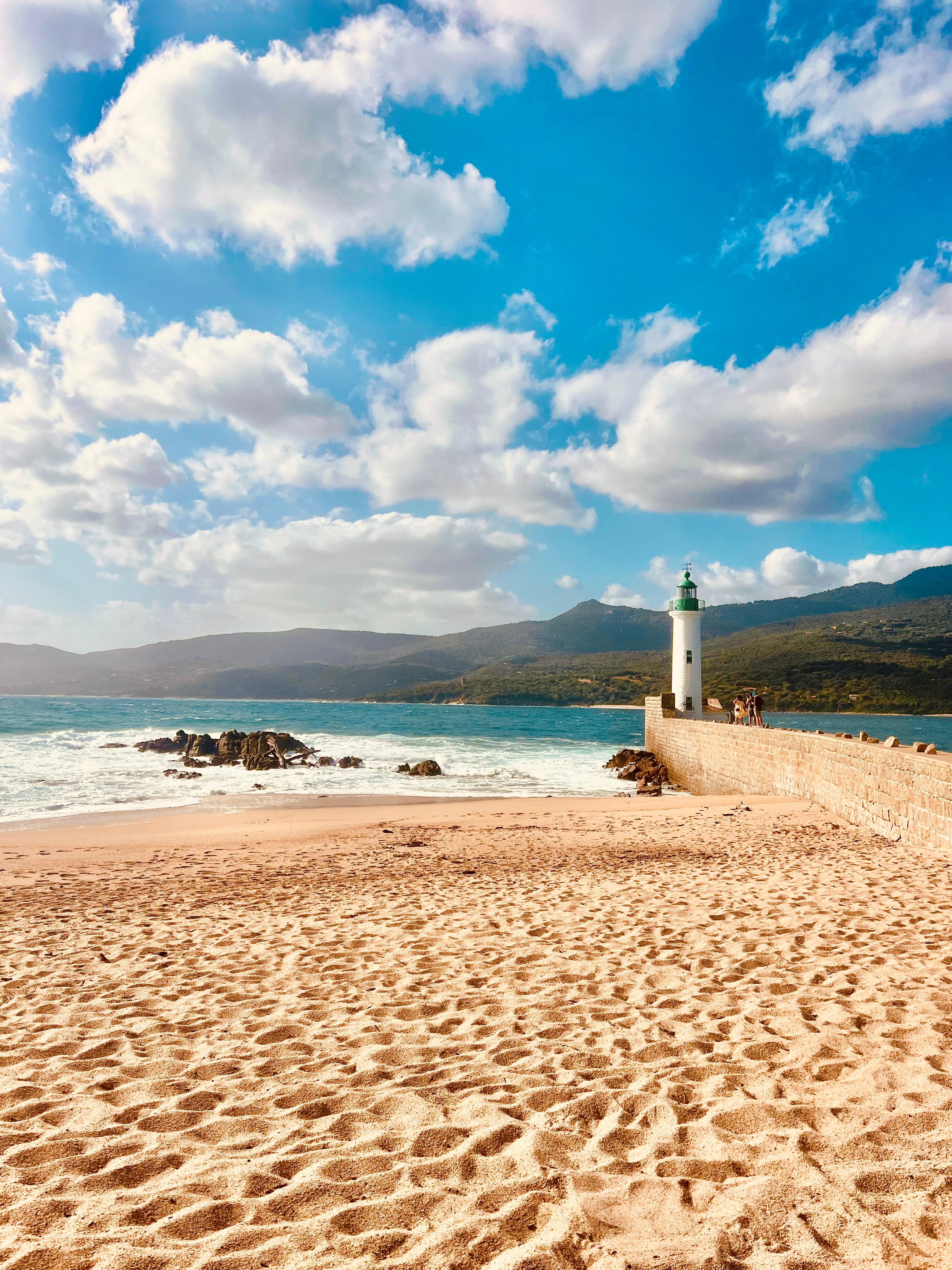 A tranquil beach scene with a lighthouse and mountains under a blue sky.