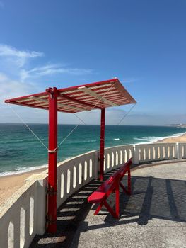 A vibrant red gazebo provides shade on a sunny beach overlooking blue ocean waves.