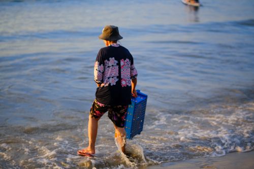 Free A man carrying a blue crate walks on My Khe Beach, Da Nang, during sunrise. Stock Photo