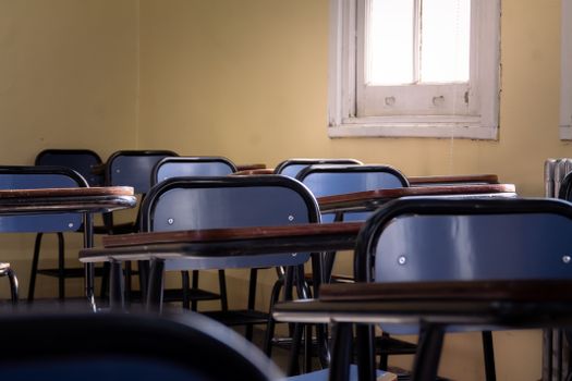 Row of empty blue chairs and wooden desks in a sunlit classroom with yellow walls.