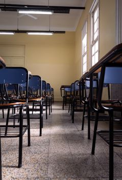 Empty classroom with neat rows of blue desks and chairs, with natural light through windows.