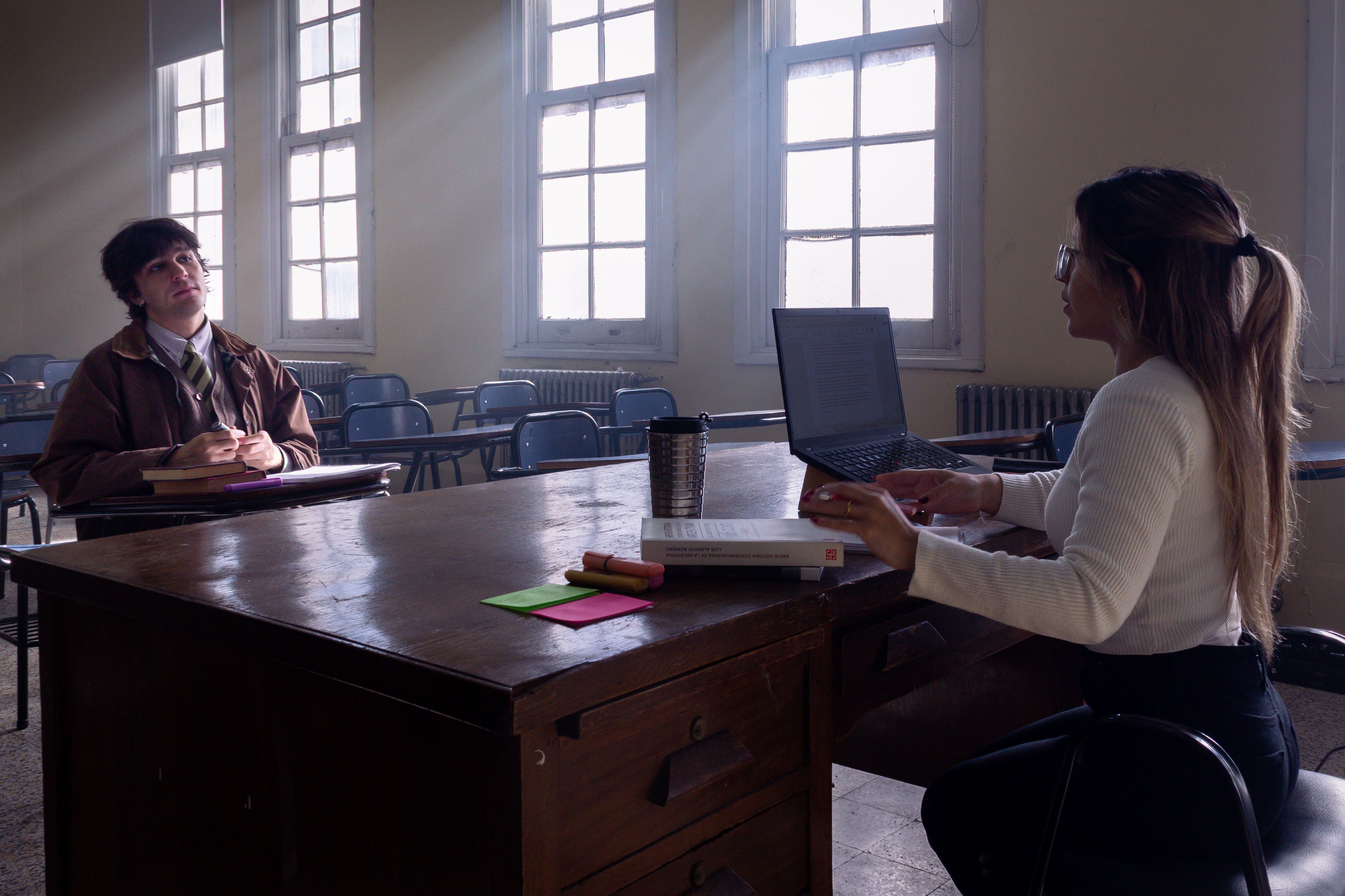 Two young adults studying in a sunlit classroom with laptops and notebooks.